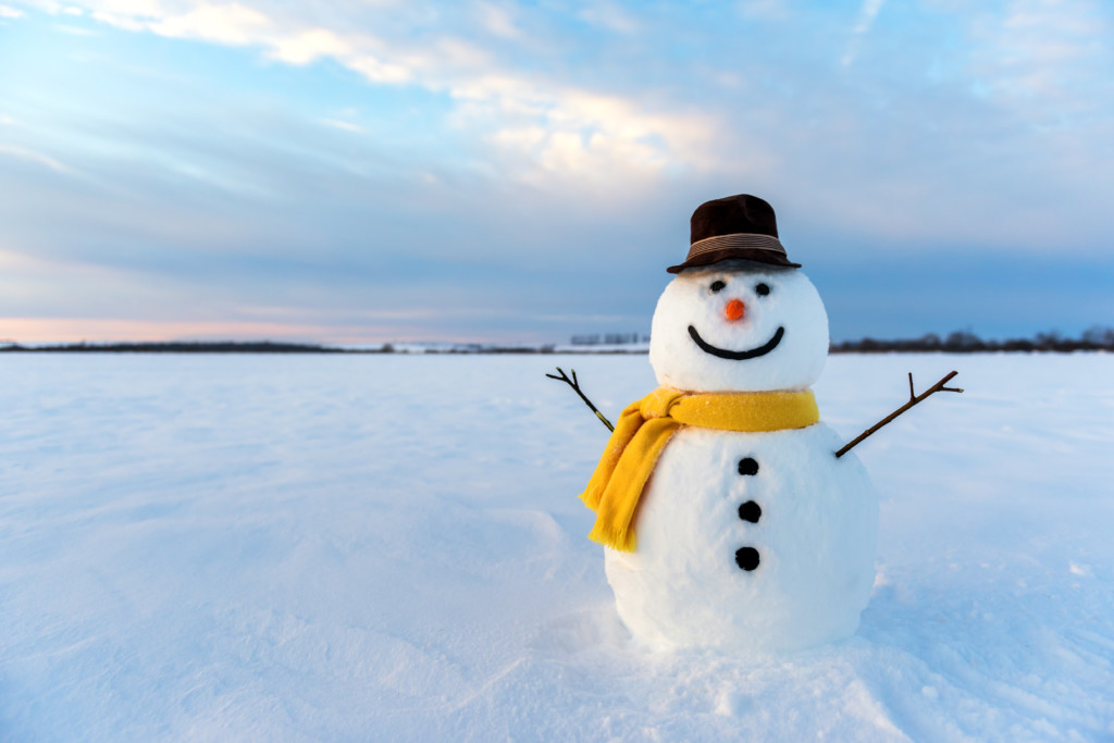 smiling snowman wearing yellow scarf & hat in a snowy field with a cloudy sunset on a winter day