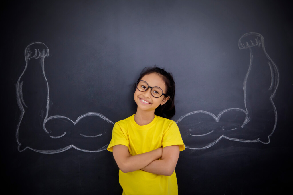 child in yellow shirt standing in front of blackboard with chalk drawing of strong muscles to emphasize being healthy & strong