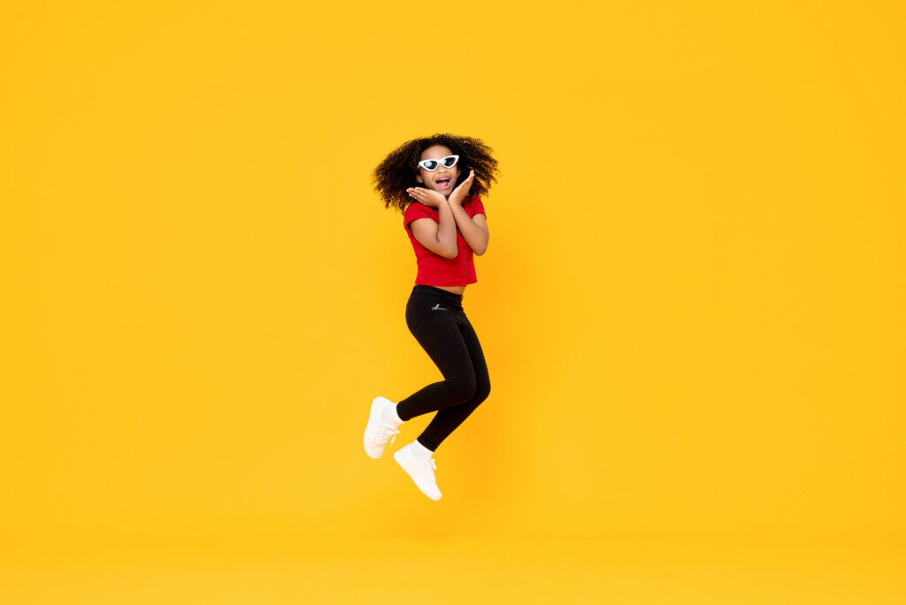 Girl in black pants red shirt & white shoes jumping in opaque yellow background listening to summer songs