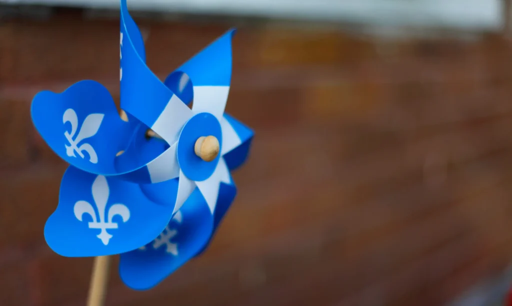 A blue and white pinwheel decorated with the Quebec fleur-de-lis symbol, held up against a soft-focus brick background. Un moulin à vent bleu et blanc orné de fleurs de lys, symbole de la culture québécoise, sur un fond de briques flou.