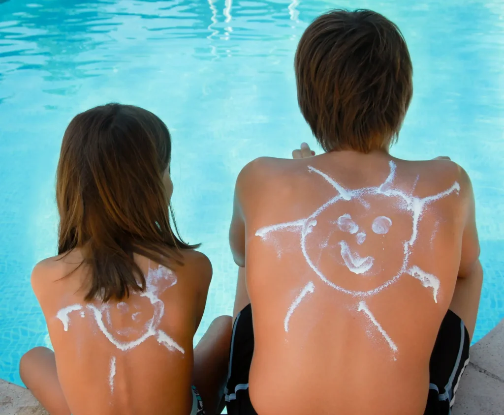 Two children sitting by a swimming pool with sunscreen art in the shape of smiling suns drawn on their backs, illustrating sun protection and summer safety Deux enfants assis au bord d'une piscine avec des dessins de soleils souriants faits en crème solaire sur leur dos, illustrant la protection solaire. chaleur