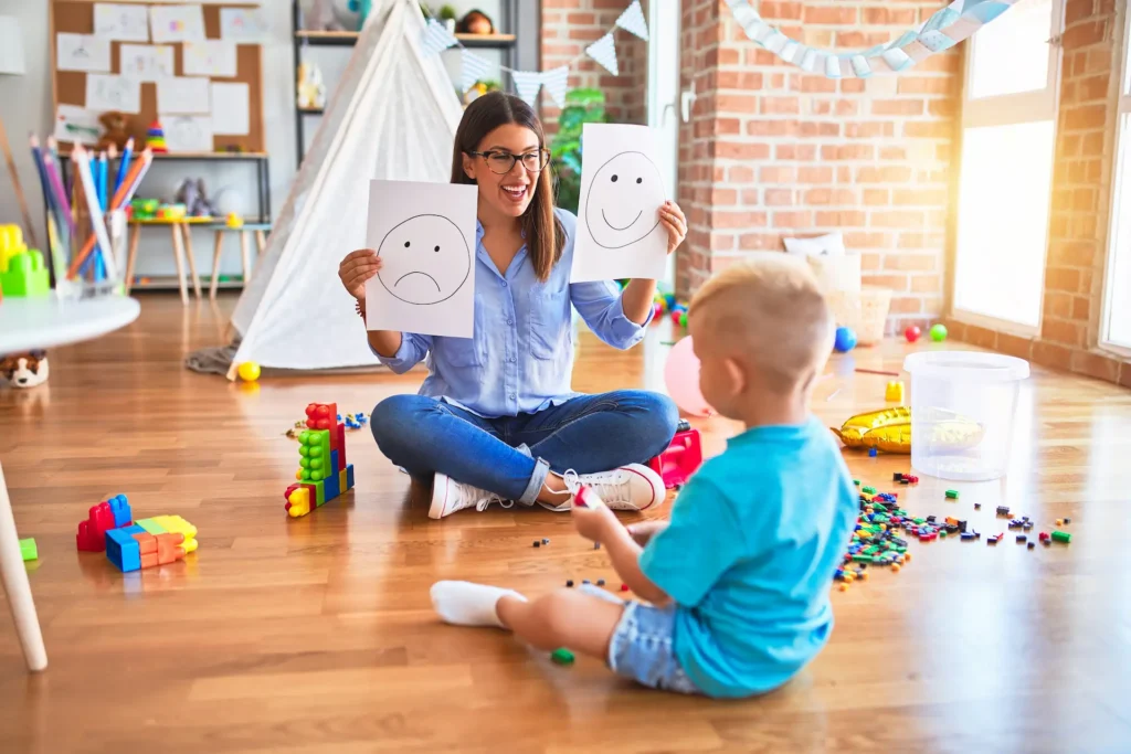 A supportive teacher or counselor sitting on the floor with a young child, holding up two drawings of faces—one sad and one happy—to help identify and discuss emotions. Une intervenante assise au sol avec un jeune enfant, tenant deux dessins représentant un visage triste et un visage joyeux pour aider à identifier et exprimer les émotions. anxiety mcgill l'anxiété