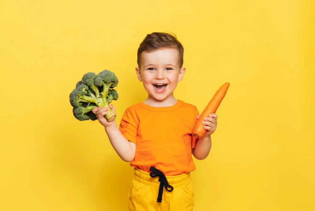 A happy young boy in an orange shirt laughing while holding a stalk of broccoli and a fresh carrot against a bright yellow background. Un jeune garçon joyeux en chandail orange riant tout en tenant un brocoli et une carotte fraîche sur un fond jaune vif. lunch