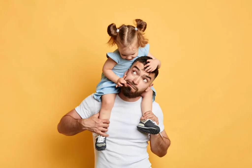 A playful young girl sitting on her father's shoulders and pulling at his face while he makes a funny expression against a bright yellow background. Une petite fille assise sur les épaules de son père et lui tirant le visage alors qu'il fait une grimace rigolote sur un fond jaune vif. Rethinking discipline