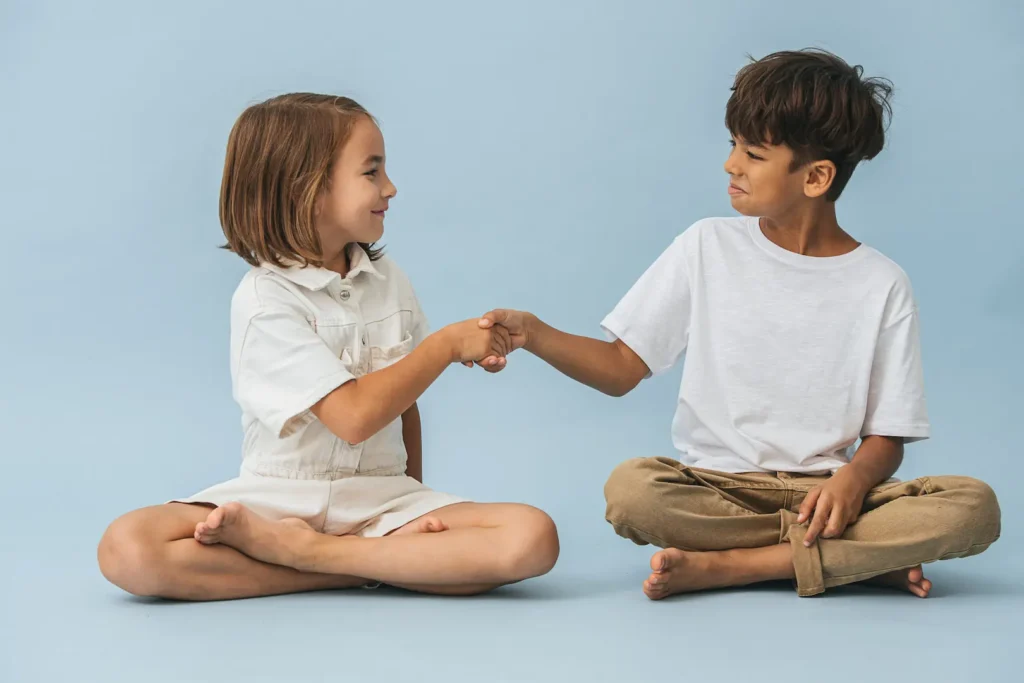 Two young children sitting cross-legged on a blue background, smiling at each other while shaking hands as a sign of friendship. Deux jeunes enfants assis par terre sur un fond bleu, se souriant tout en se serrant la main en signe d'amitié. Friendship