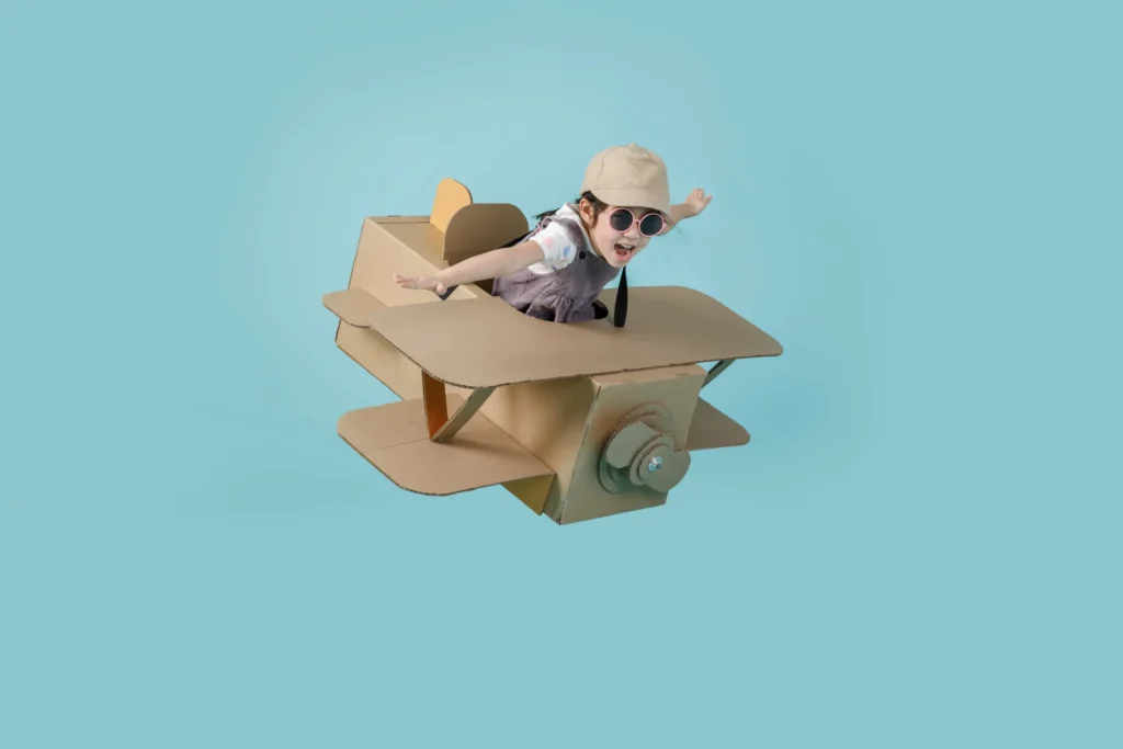 A young girl wearing a cap and sunglasses, excitedly playing in a DIY airplane made from cardboard boxes against a light blue background. Une jeune fille portant une casquette et des lunettes de soleil, s'amusant dans un avion artisanal fabriqué en carton sur un fond bleu clair. Confidence. Confiance