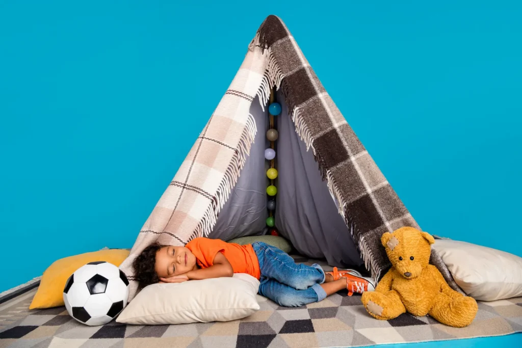 A young child sleeping peacefully inside a cozy blanket tent decorated with lights, surrounded by a soccer ball and a teddy bear against a blue background. Un jeune enfant dormant paisiblement dans une tente de couvertures décorée de lumières, entouré d'un ballon de soccer et d'un toutou sur un fond bleu. Bedtime coucher