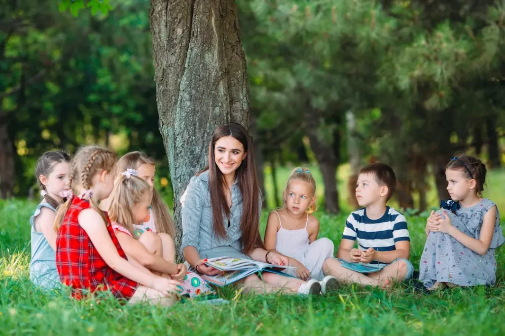 A teacher sitting on the grass in a park, reading a book to a diverse group of attentive young children during an outdoor lesson. Une enseignante assise sur l'herbe dans un parc, lisant un livre à un groupe d'enfants attentifs lors d'une leçon à l'extérieur.
