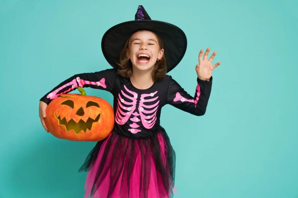 A young girl laughing joyfully while wearing a pink and black skeleton costume and a witch hat, holding a carved pumpkin against a turquoise background. Une jeune fille riant joyeusement en costume de squelette rose et noir avec un chapeau de sorcière, tenant une citrouille sur un fond turquoise. Thrift Friperie