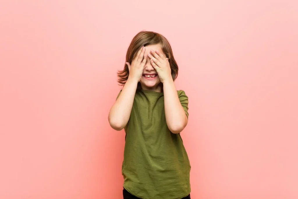 A young child in a green t-shirt playfully covering their eyes with both hands against a solid pink background, representing privacy and anonymity. Un jeune enfant en t-shirt vert se couvrant les yeux avec les mains sur un fond rose, illustrant le concept de vie privée et d'anonymat. Digital privacy privée