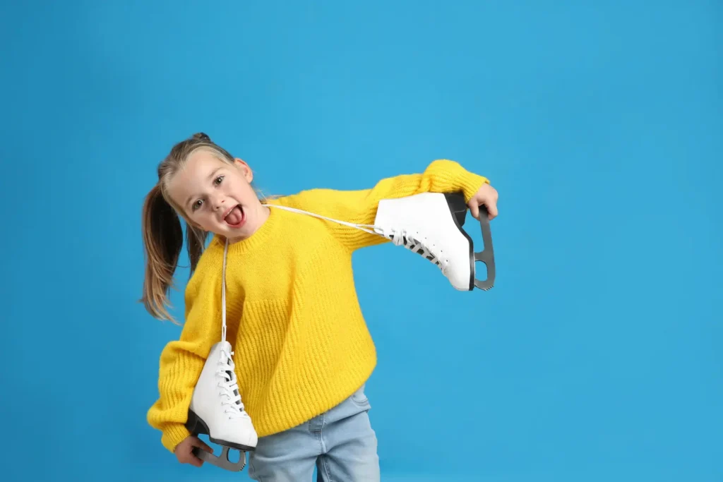 A playful young girl in a bright yellow sweater holding a pair of white figure skates over her shoulders against a blue background, looking excited. Une jeune fille enjouée portant un chandail jaune vif et tenant une paire de patins à glace blancs sur ses épaules sur un fond bleu. Life skill Compétences de vie