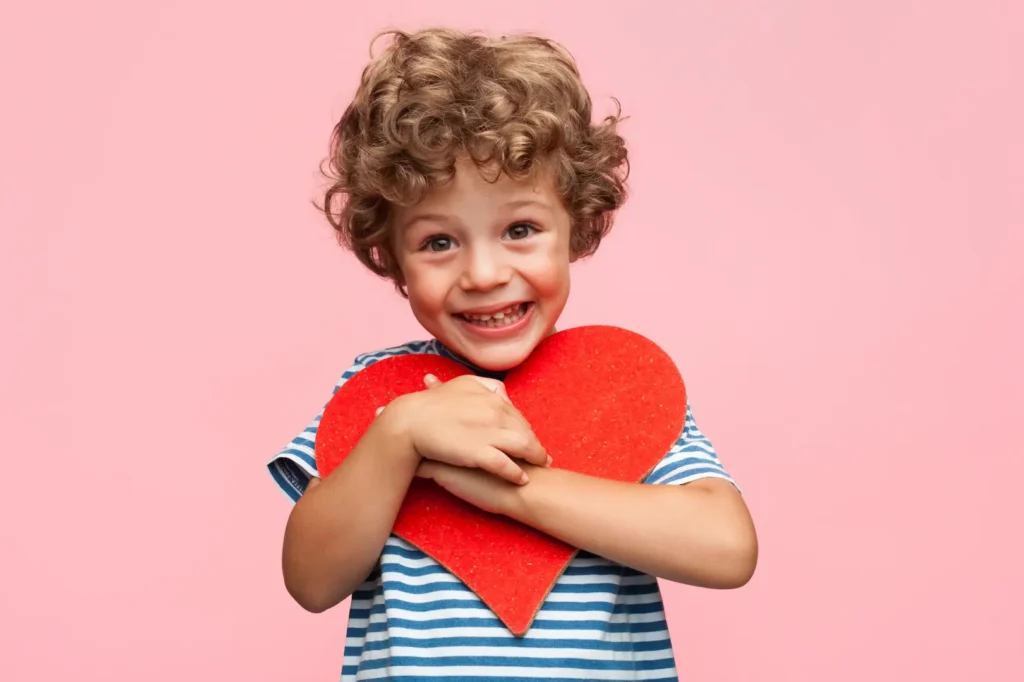 A smiling young boy with curly hair happily hugging a large red heart-shaped cutout against a solid pink background. Un jeune garçon souriant aux cheveux bouclés serrant joyeusement un grand cœur rouge en carton contre lui sur un fond rose. Help aider