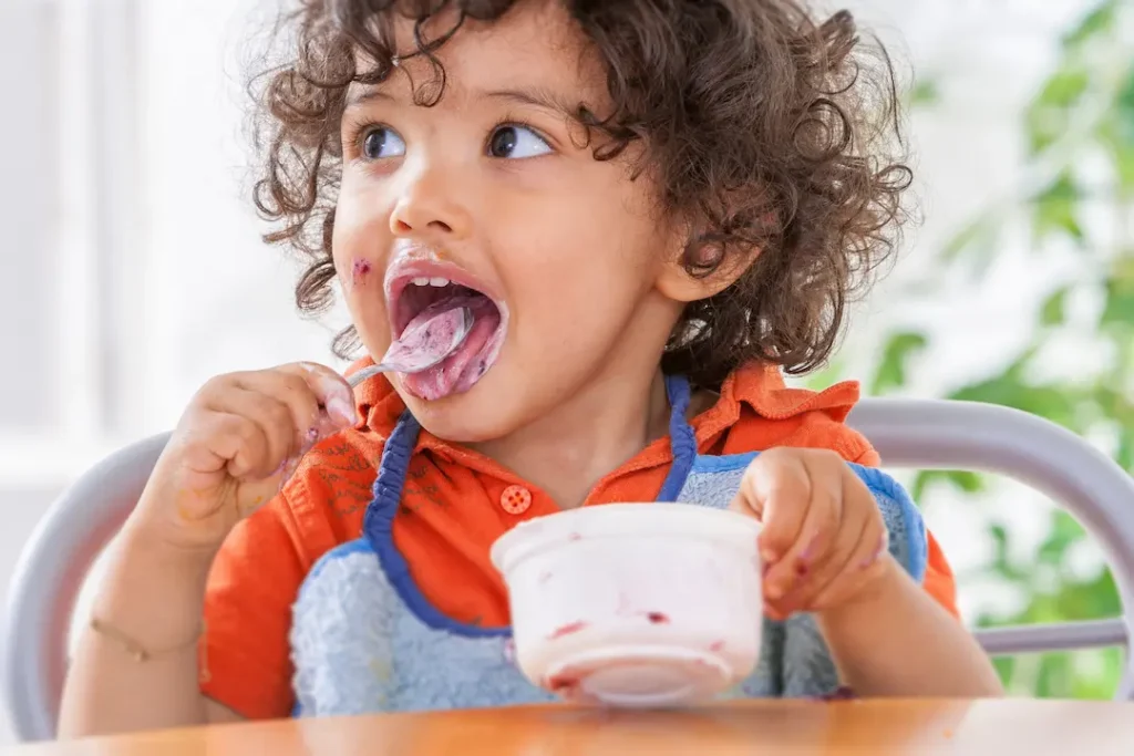 A young child with curly hair and an orange shirt happily eating yogurt with a spoon, showing a joyful and healthy relationship with food. Un jeune enfant aux cheveux bouclés portant un chandail orange mangeant joyeusement du yogourt, illustrant un rapport positif à l'alimentation. nourriture