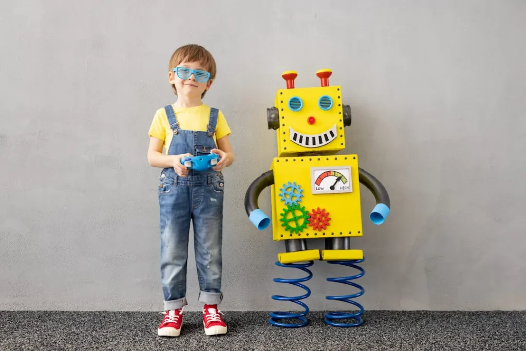 A young boy in overalls and blue shutter shades holding a remote control next to a friendly, homemade yellow cardboard robot on blue spring legs. Un jeune garçon en salopette tenant une télécommande à côté d'un robot artisanal jaune en carton, illustrant l'apprentissage des technologies. Critical thinking pensée critique