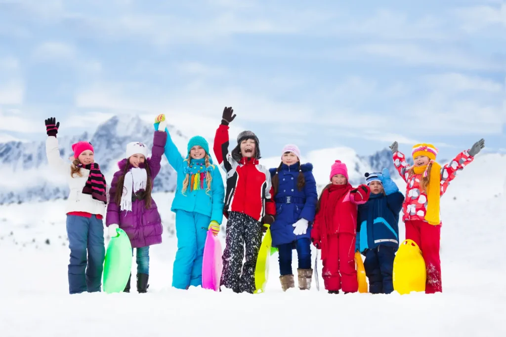 A group of cheerful children in colorful winter gear standing in the snow with sleds, waving their hands joyfully against a mountain backdrop. Un groupe d'enfants joyeux en vêtements d'hiver colorés debout dans la neige avec des luges, levant les mains avec enthousiasme devant des montagnes. Movement mouvement