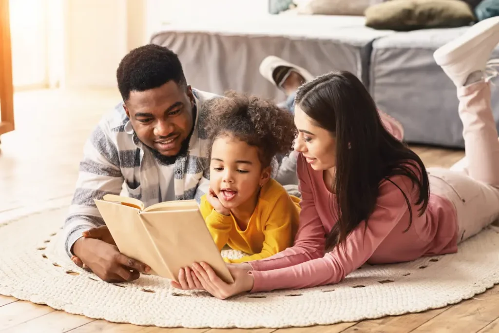 A smiling family of three—a father, mother, and young daughter—lying together on a round rug at home, focused on reading a book together to illustrate the value of shared quality time. Une famille souriante composée d'un père, d'une mère et d'une jeune fille, allongés ensemble sur un tapis rond à la maison pour lire un livre, illustrant la valeur du temps de qualité partagé.
