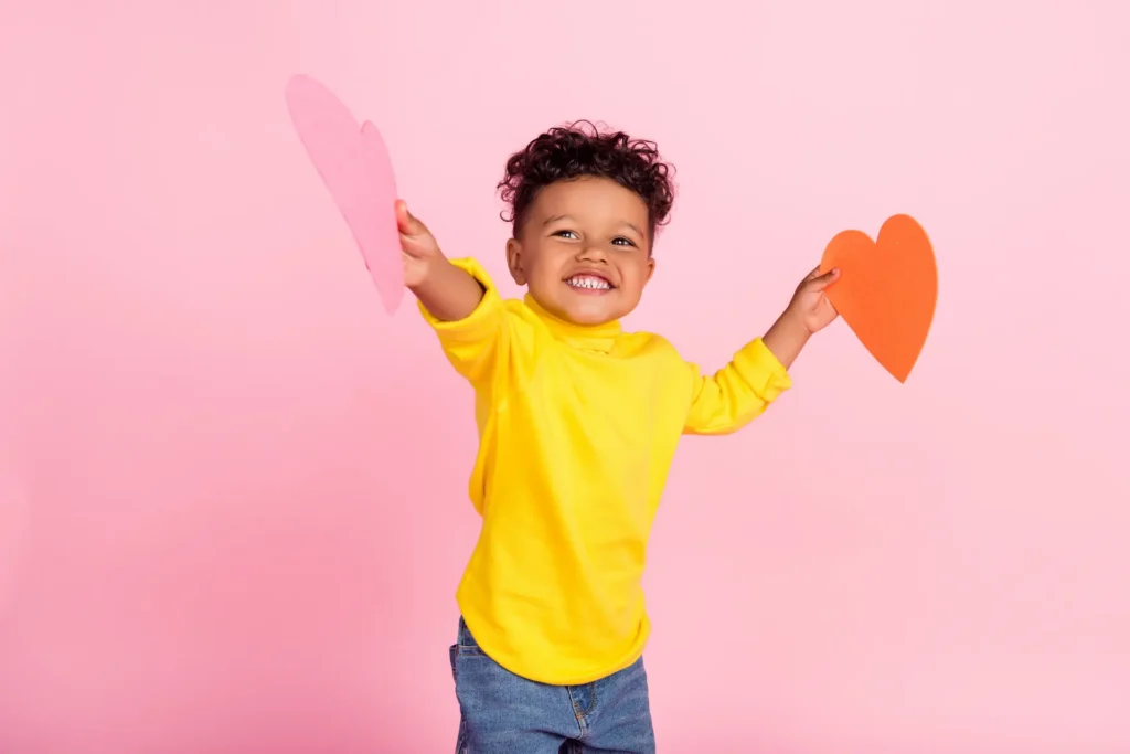 A joyful young boy in a bright yellow sweater holding up two large paper hearts against a pink background, expressing the excitement and love associated with holiday celebrations. Un jeune garçon joyeux portant un chandail jaune vif et tenant deux grands cœurs en papier sur un fond rose, exprimant le bonheur et l'amour associés aux célébrations festives. Fetes