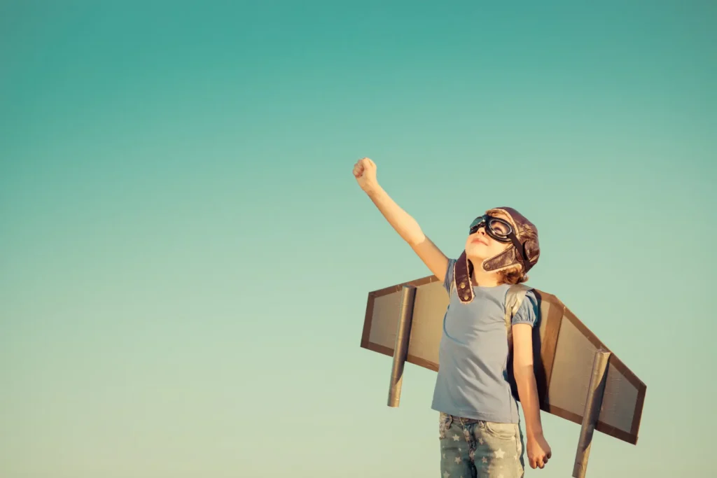 A young child wearing a vintage aviator hat, goggles, and cardboard wings, reaching one fist toward the clear blue sky in a superhero pose, representing limitless imagination and gentle aspiration. Un jeune enfant portant un casque d'aviateur vintage, des lunettes de protection et des ailes en carton, levant le poing vers le ciel bleu dans une pose de super-héros, représentant l'imagination débordante et l'aspiration sereine. Goal Objectif