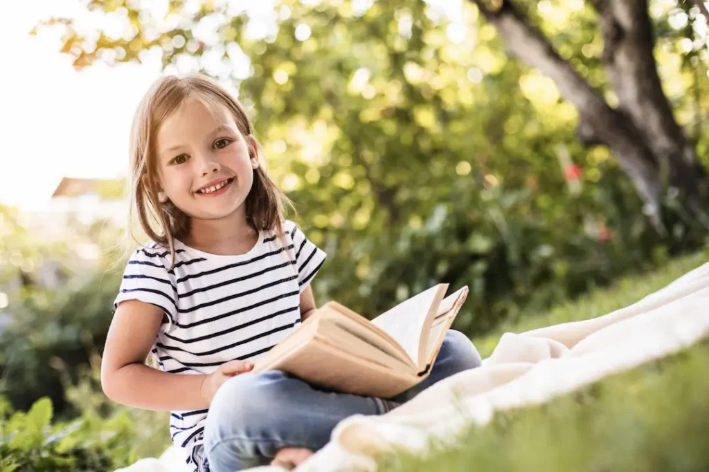 A young girl sitting on a blanket in a sunlit garden, smiling while reading a book, representing the joy of discovery and the foundation of lifelong learning. Une jeune fille assise sur une couverture dans un jardin ensoleillé, souriante avec un livre ouvert, illustrant le plaisir de la découverte et la base de l'apprentissage continu. lecture