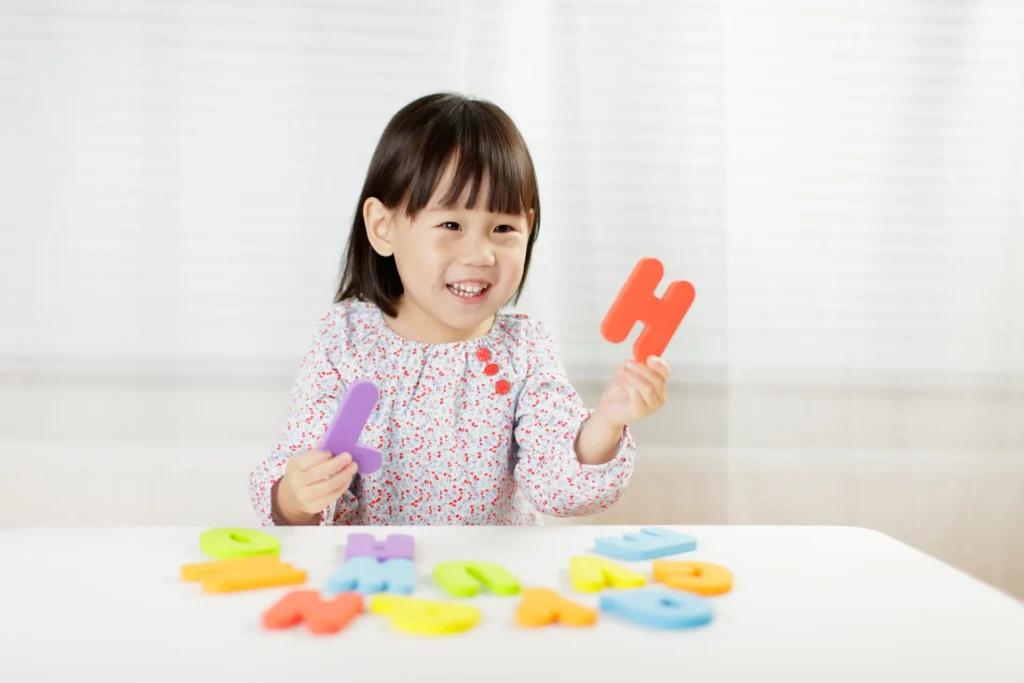 A smiling young girl sitting at a white table, happily playing with large, colorful foam letters to build words and practice her alphabet skills by playing a game. Une jeune fille souriante assise à une table blanche, s'amusant joyeusement avec de grandes lettres en mousse colorées pour former des mots et pratiquer l'alphabet avec l'aide des jeux.