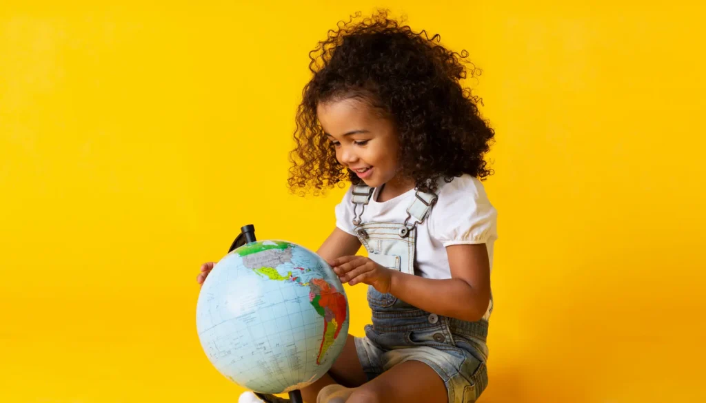 A young girl with curly hair smiling as she explores a world globe against a bright yellow background, representing global mindedness and the discovery of diverse cultures. Une jeune fille aux cheveux bouclés souriant en explorant un globe terrestre sur un fond jaune vif, illustrant la curiosité et la découverte de nouvelles cultures. monde Gift cadeau