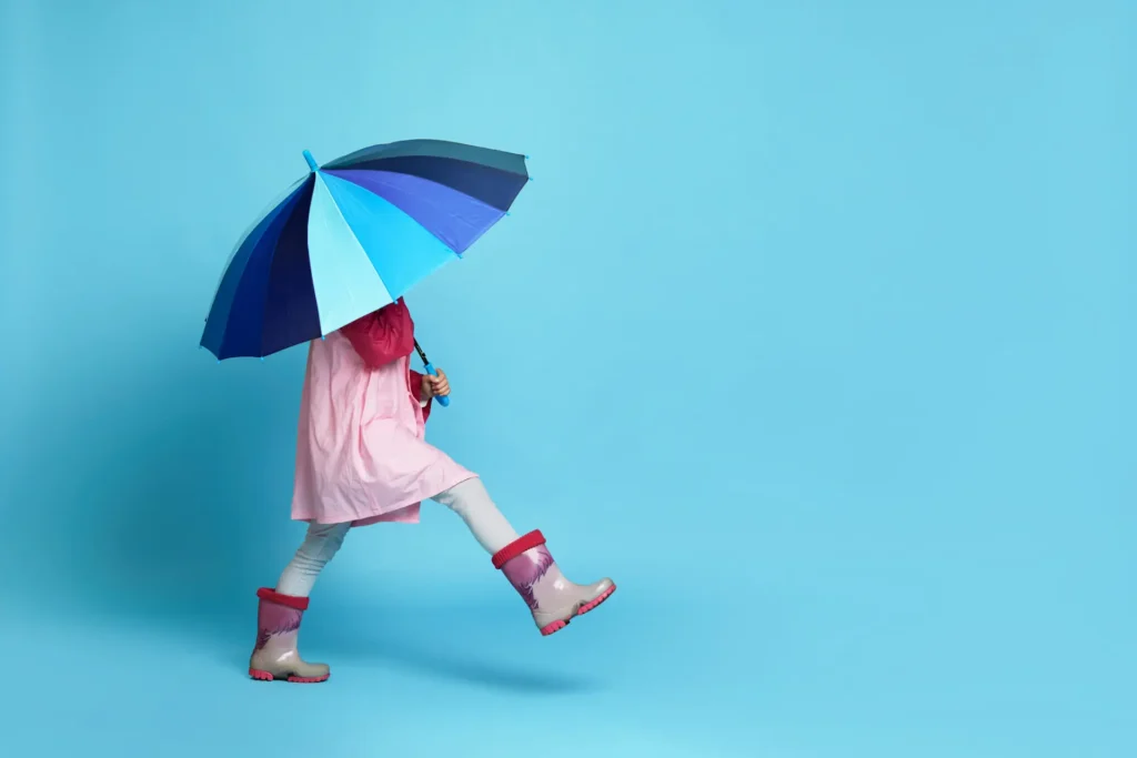 A child in a pink raincoat and boots walking playfully while holding a large, colorful umbrella that hides their face, representing the need for protection and a safe space during difficult moments. Un enfant en imperméable rose et bottes de pluie marchant joyeusement tout en tenant un grand parapluie coloré qui cache son visage, illustrant le besoin de protection et de sécurité lors de moments difficiles.