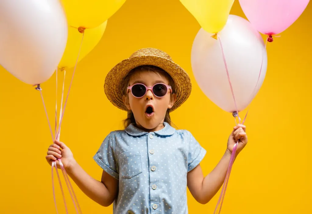 A young girl with an amazed expression, wearing a straw hat and pink sunglasses, holding a bunch of white and yellow balloons against a bright yellow background, capturing the excitement of the new Plateau summer camp location. Une jeune fille à l'expression émerveillée, portant un chapeau de paille et des lunettes de soleil roses, tenant des ballons blancs et jaunes sur un fond jaune vif, illustrant l'excitation entourant notre nouvel emplacement sur le Plateau.