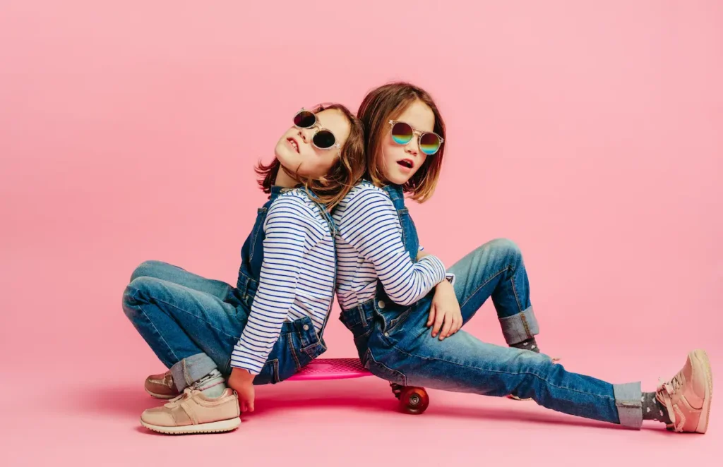 Two young girls sitting back-to-back on a pink skateboard against a solid pink background, wearing matching striped shirts, denim overalls, and sunglasses, representing the fun and social connection of summer activities. Deux jeunes filles assises dos à dos sur une planche à roulettes rose sur un fond rose, portant des salopettes en jean et des lunettes de soleil, illustrant le plaisir et la camaraderie des activités d'été. Languages langue
