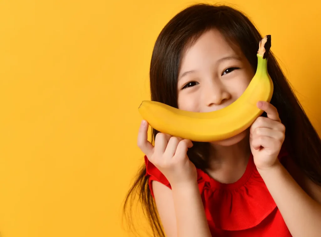 A smiling young girl holding a yellow banana like a grin across her face against a bright yellow background, representing healthy nutrition and the presence of lectins in common fruits. Une jeune fille souriante tenant une banane jaune devant sa bouche comme un sourire sur un fond jaune vif, illustrant la nutrition saine et la présence de lectines dans les fruits communs.