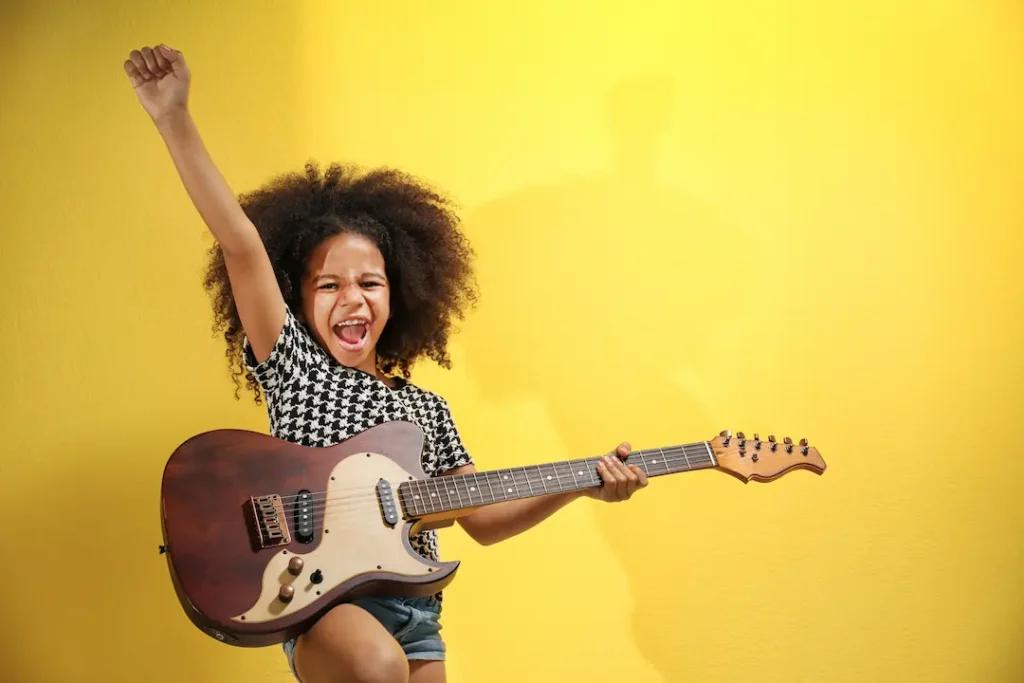 A young girl with curly hair joyfully shouting and holding an electric guitar with one arm raised in a victory pose against a bright yellow background, representing the bold confidence built through creative expression and music with confidence. Une jeune fille aux cheveux bouclés criant de joie et tenant une guitare électrique avec un bras levé en signe de victoire sur un fond jaune vif, illustrant la confiance en soi par l'expression créative.