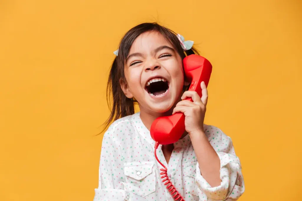 A young girl laughing joyfully while holding a bright red vintage telephone receiver to her ear against a yellow background, representing effective communication and the excitement of connection and skills. Une jeune fille riant joyeusement tout en tenant un combiné de téléphone rouge vintage à son oreille sur un fond jaune, illustrant une communication efficace et le plaisir de la connexion. Compétences