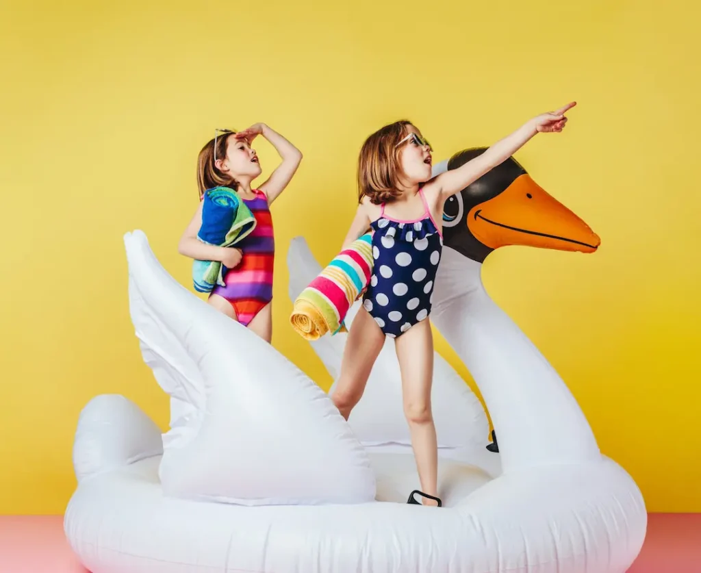 Two young girls in swimsuits holding rolled-up towels and standing on a large white swan pool float against a yellow background, capturing the excitement and adventurous spirit of summer camps . Deux jeunes filles en maillot de bain tenant des serviettes roulées et debout sur un grand cygne gonflable sur un fond jaune, capturant l'excitation et l'esprit d'aventure des camps d'été.