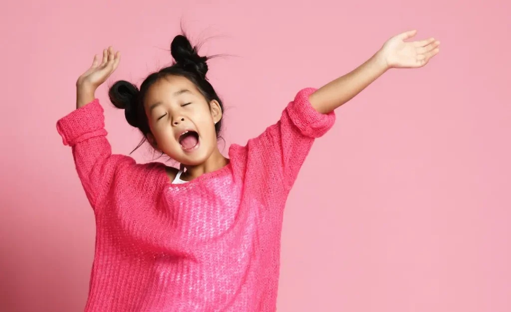 A young girl with her hair in two buns, eyes closed and mouth open in a big yawn while stretching her arms, perfectly capturing a moment of boredom or tiredness against a bright pink background. Une jeune fille aux cheveux coiffés en deux macarons, les yeux fermés et la bouche grande ouverte dans un bâillement tout en étirant les bras, illustrant un moment d'ennui ou de fatigue sur un fond rose vif.