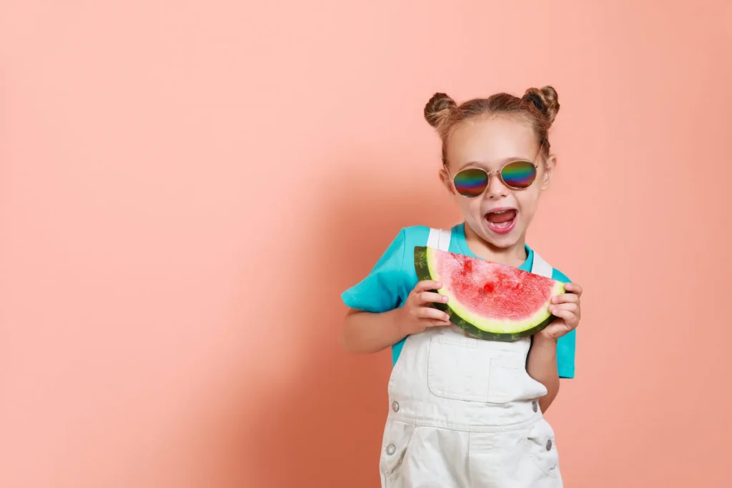 A young girl with space buns and colorful sunglasses holding a large slice of watermelon and smiling excitedly against a peach-colored background. Summer été
