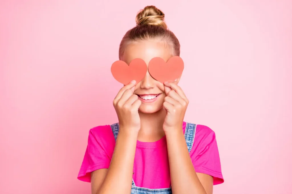 A smiling young girl holding two red paper hearts over her eyes against a pink background, representing the joy and playfulness of celebrating love and connection. Une jeune fille souriante tenant deux cœurs en papier rouge sur ses yeux sur un fond rose, illustrant la joie et le plaisir de célébrer les liens affectifs. amour