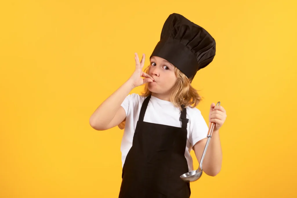 A young child dressed in a black chef's hat and apron, making a 